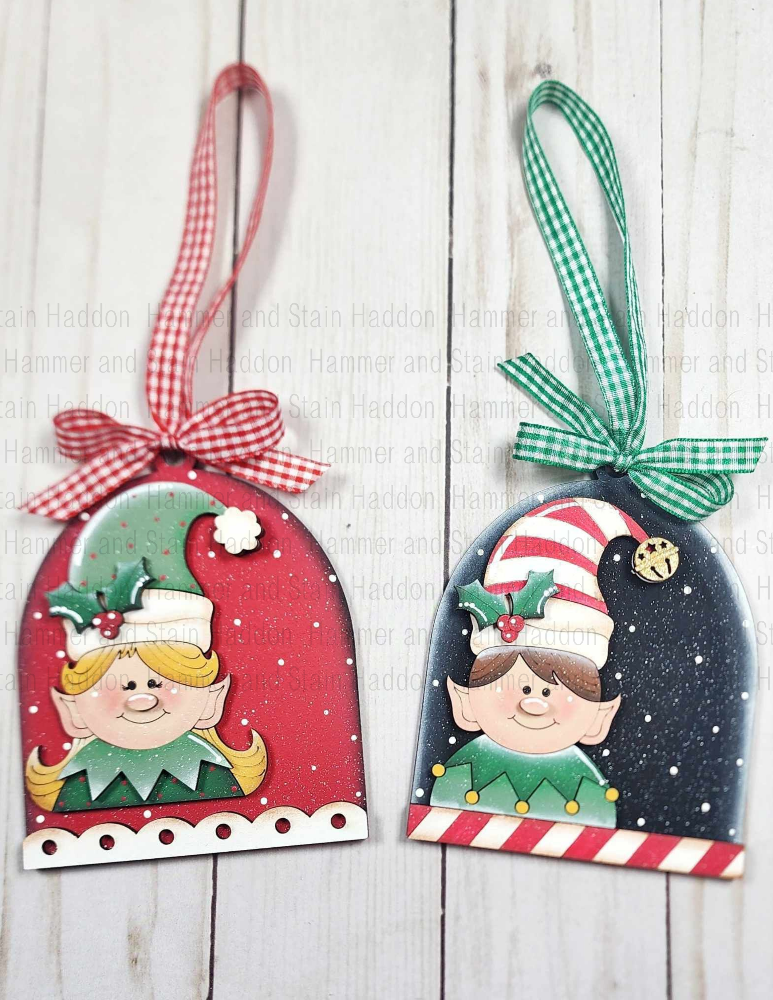 Two Christmas-themed elf ornaments with colorful bows on a wooden surface.