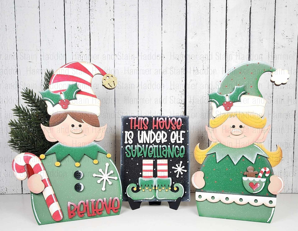 Two Christmas-themed elf decorations and a sign with text on a wooden background.