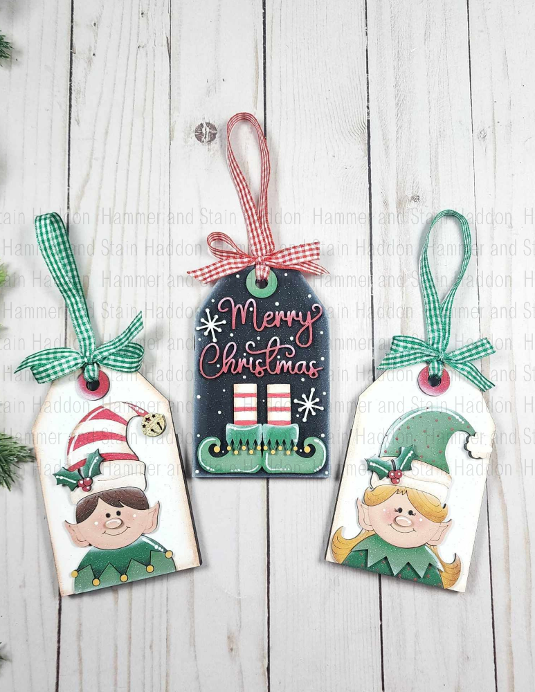 Three Christmas-themed gift tags with elves and 'Merry Christmas' text on a wooden surface.