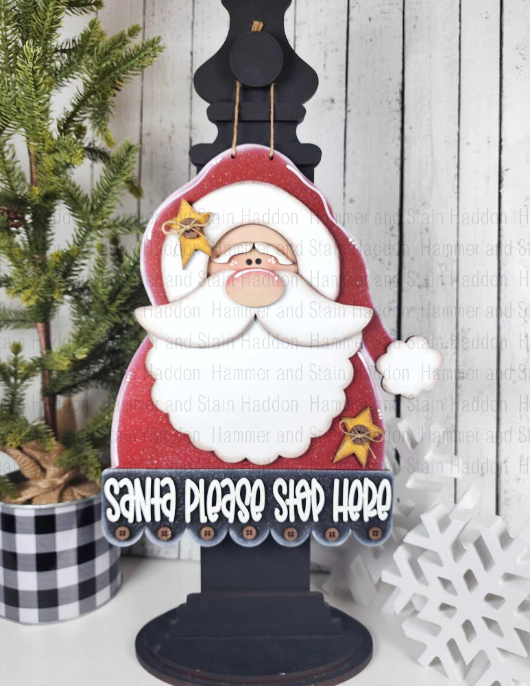 Decorative sign with Santa Claus and text 'Santa Please Stop Here' on a wooden stand, Christmas tree and snowflake decorations in the background.