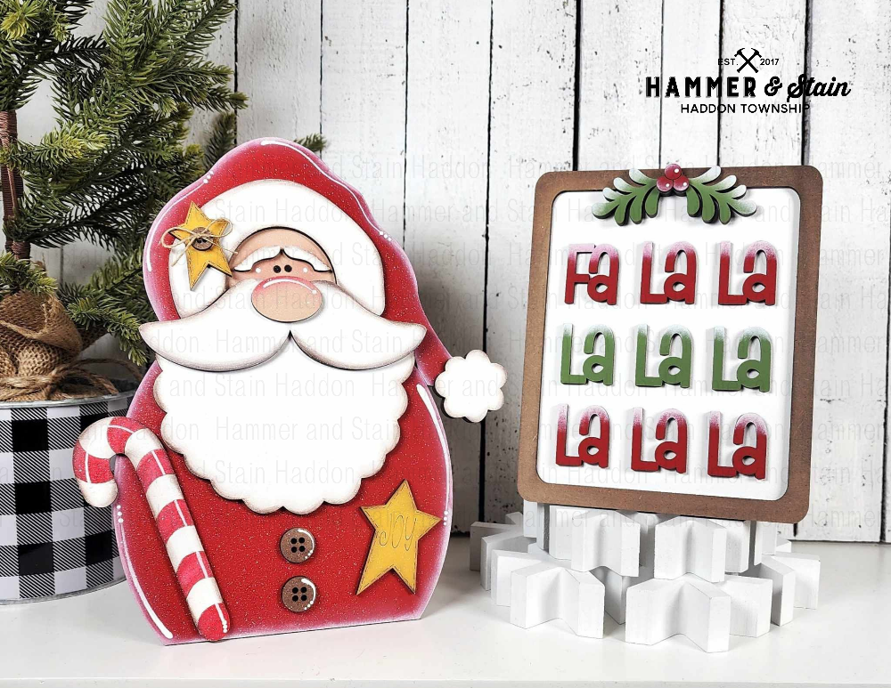 Decorative Santa Claus shelf sitter with candy cane and star, next to a sign with 'Fa La La' text, on a wooden background.