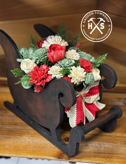 Decorative sleigh with wood flowers and ribbons on a wooden surface, featuring the Hammer and Stain logo.